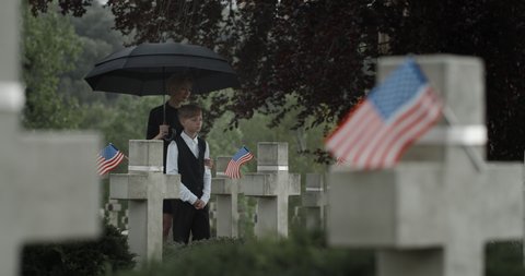 Mother and her teen son under umbrella putting flowers at cemetery. Widow and child walking and stopping near gravestone. Concept of memorial day. American flags on stone crosses.