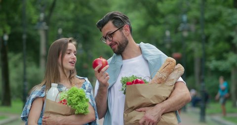 Good looking couple coming back from grocerie shop and holding paper bags full of food. Man in glasses throwing pomegranate up while walking through park with his pretty wife.