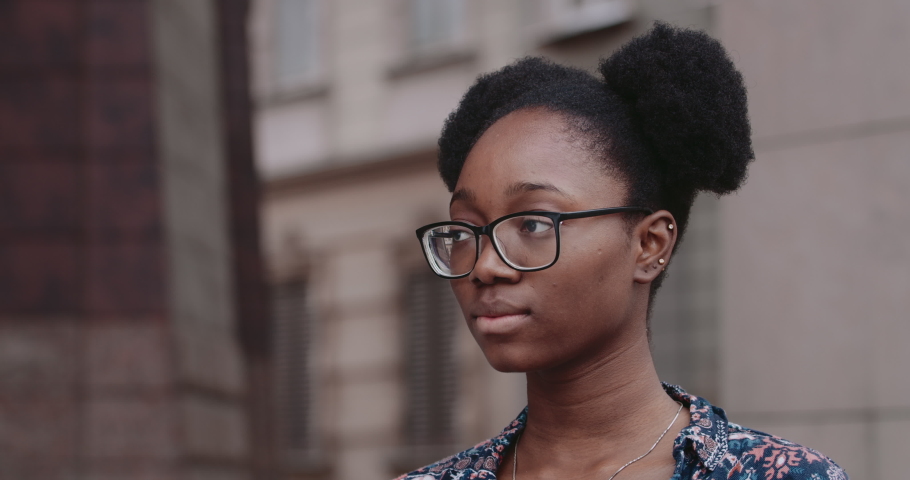 Close up view of afro american woman in glasses turning head and looking to camera. Portrait of serious millennial girl standing at city street.Concept of people and emotions. | Shutterstock HD Video #1056537326