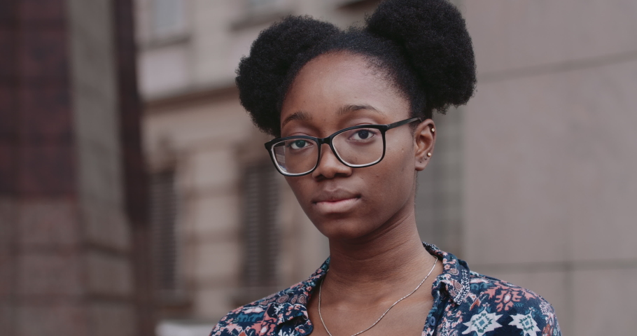 Close up view of afro american woman in glasses turning head and looking to camera. Portrait of serious millennial girl standing at city street.Concept of people and emotions. Royalty-Free Stock Footage #1056537326