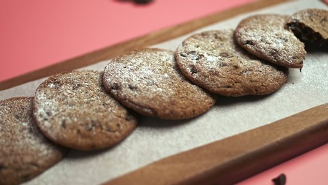 Freshly made Chocolate Chip Cookies on a wooden serving board. Cookies with a glass of fresh milk on the side along with chocolate chips and pieces of chocolate around on a pink background.
