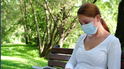 Young woman sits alone in a Park in a mask and reads a book during the epidemic. Safety, distance, health, N1H1, virus protection, covid, virus and coronavirus concept