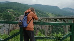 Back view of Young Woman Taking Pictures of Djurdjevic bridge. Hiker Girl taking photos or video with digital camera of beautiful canyon landscape while travel in Montenegro.
