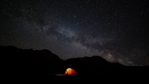 Orange tent with a burning light inside stands in a clearing under the starry sky. On the horizon mountains alps. Concept of starry night sky and Milky Way. camping in the mountain. timelapse video