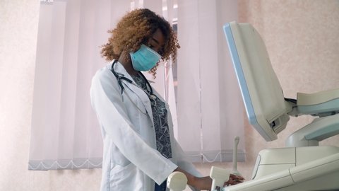 Close-up of face of professional african american obstetrician gynecologist in protective mask performing ultrasound of belly of pregnant woman. Concept of medicine, health care and people