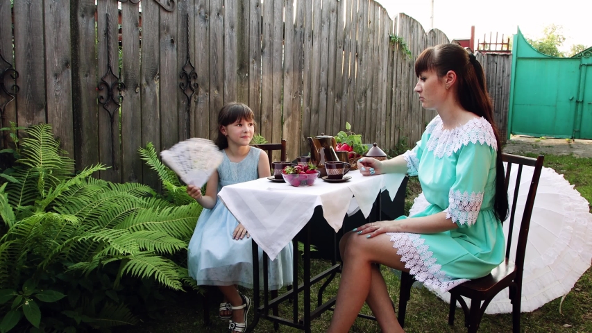 A little girl sits at a table with her mother in the yard. She fanns herself as mom pours tea into a cup. Young ladies in beautiful dresses. Summer day.
