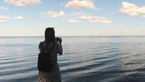 Rear view portrait of a woman making selfie in the background of a beautiful sea landscape with smartphone and monopod at sunset