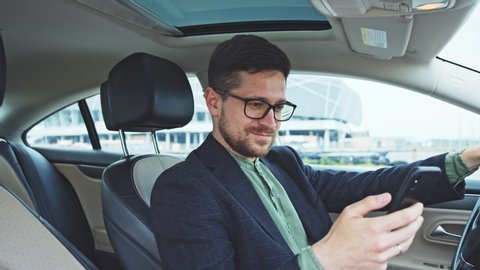 Joyful bearded young entrepreneur enjoying successful life sitting in expensive car and browsing modern smartphone. Business people. Prosperity.