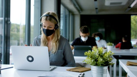 Young woman with face mask back at work in office after lockdown, having video call.
