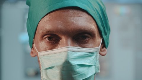 Closeup of tired experienced male doctor removes protective mask from his face and breath gulp of fresh air after many operations performed on his shift in hospital during COVID-19 Coronavirus attack