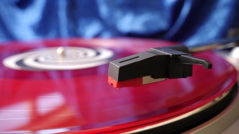 Stylus arm and needle lowered on 12” red vinyl record on a DJ turntable on blue velvet background. Retro LP platter. Close up. Hi-fi musical equipment.  