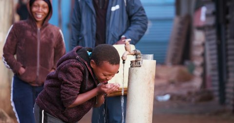 Poverty.Inequality.Water crisis. Close-up view of a young black african boy drinking water from a communal tap while people line up to collect water in plastic containers 