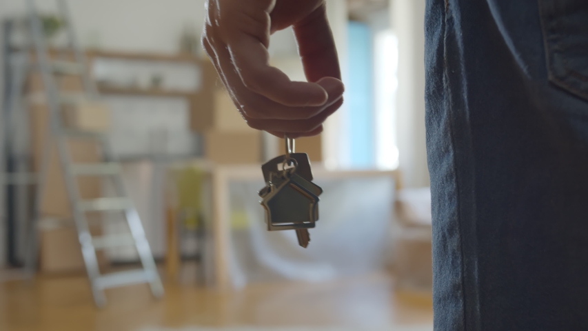 Close up of man holding key with house keychain in hand and walking in new apartment with cardboard boxes and ladder on background. Guy moving in new home. Buying or renting real estate concept Royalty-Free Stock Footage #1056568946