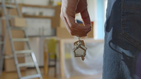 Close up of man holding key with house keychain in hand and walking in new apartment with cardboard boxes and ladder on background. Guy moving in new home. Buying or renting real estate concept