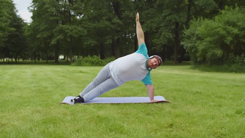 Portrait of stout man in headphones doing yoga exercises on fitness mat outdoors. Overweight guy losing weight and working out in summer park. Healthy lifestyle, sport concept