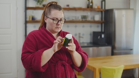 Overweigh woman reading instructions on bottle of herbal extract natural diet pills. Young fat girl in bathrobe with slimming pills standing in modern loft apartment