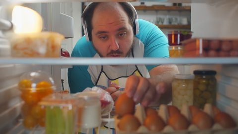 View from fridge of cheerful overweight man in apron and headphones taking eggs from shelf making breakfast. Fat guy cooking dinner and taking ingredients in refrigerator
