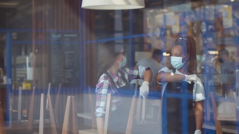 View through window of multiethnic waitresses in protective masks and gloves doing elbow bumps greeting in restaurant before opening. Covid-19 restrictions and small business crisis concept