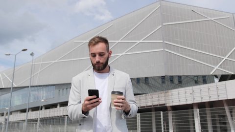Young businessman in a light suit goes with coffee and uses his phone to chat with colleagues on the background of a modern building, smiling, business partners