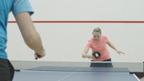 Portrait of positive sportswoman playing ping-pong with unrecognizable sportsman. Cheerful young Caucasian woman hitting ball with racket and talking. Joyful blond girl training table tennis in gym.