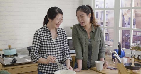 slow movement of two asian korean girl roommates standing at wooden table island in home kitchen. woman looking at friend whisking eggs and milk in bowl and talking to her. ladies relax in apartment