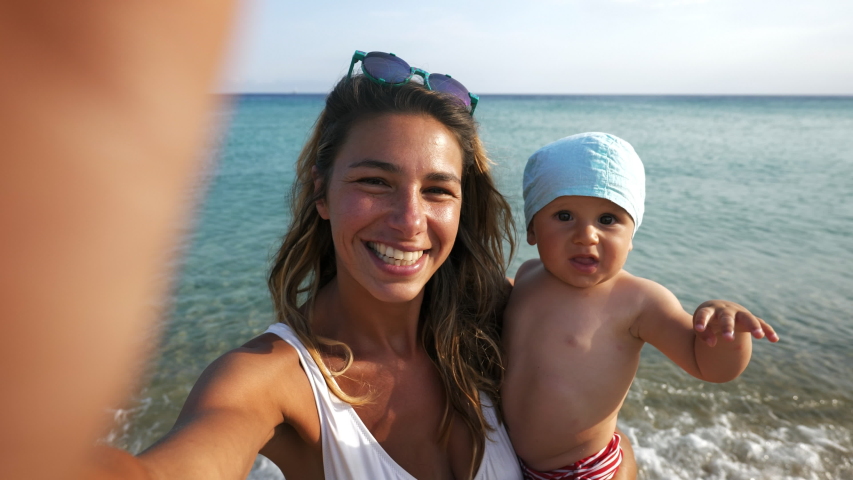 Authentic shot of happy carefree smiling neo mother and her newborn baby making selfie or video call to father or relatives on a beach with seaside in a sunny day.  Royalty-Free Stock Footage #1056581264