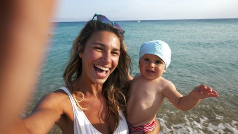 Authentic shot of happy carefree smiling neo mother and her newborn baby making selfie or video call to father or relatives on a beach with seaside in a sunny day. 