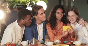 Attractive girl holding phone horizontally while showing friends funny photos. Millennial cheerful people laughing and looking at mobilephone screen while sitting in cosy cafe