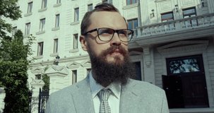 A close-up video of a bearded, nice looking, hipster businessman is looking for his business partner. He is wearing stylish glasses . An old style neoclassical building beautifies his background.
