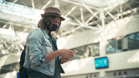 A young woman with a backpack and in a facial protective mask standing at the airport hall and using her smartphone.