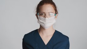 Attractive female doctor in protective eyeglasses, mask and medical gloves happily showing thumbs up gesture over white background