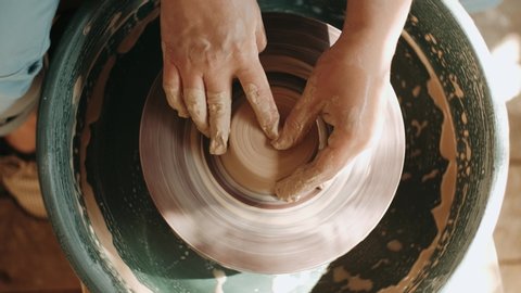 Close up of hands working clay on potter's wheel. Potter shapes the clay product with pottery tools on the potter's wheel, top view, craft factory authentic.
