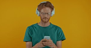 happy redhead man in headphones dancing with smartphone isolated on yellow