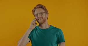 positive redhead man in glasses talking on smartphone isolated on yellow