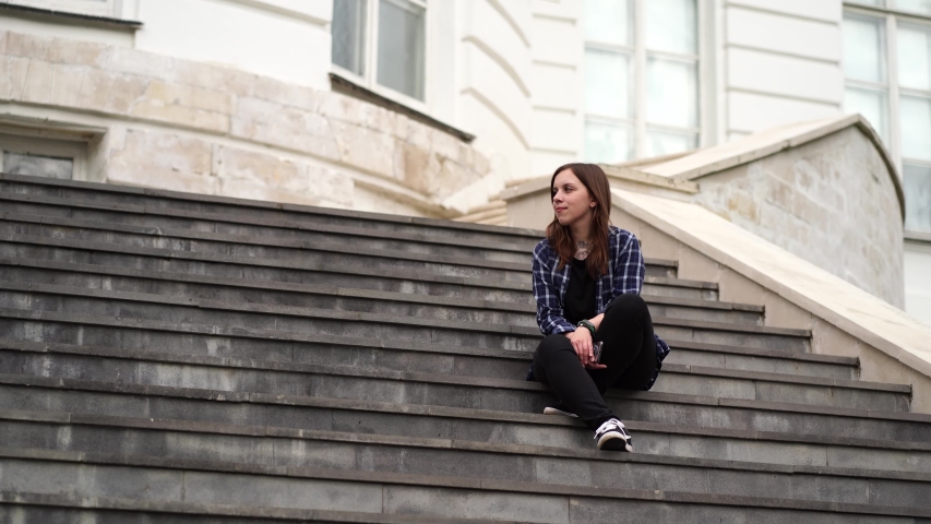 Young woman flipping news in smartphone, sitting on stairs of manor. Adult female with mobile phone sitting on steps of Palace.
