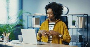 Pretty happy African American young woman sitting at office and talking on webcam on laptop in headset. Beautiful female worker having video call on computer at working place indoor. Job concept