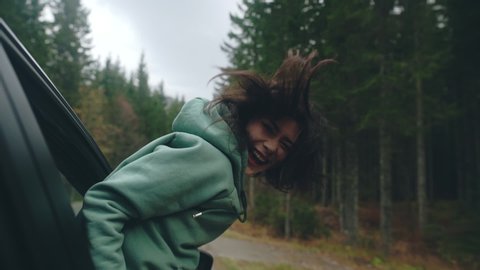 Close-up face of young woman traveling by car while road trip. Woman pulling her face and hands out of car window and enjoying speed while her hair flying away because of the wind. Concept of freedom.