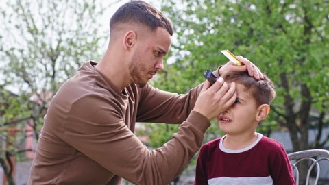 In a sunny day in the garden a little child boy his big brother make a hair cut in with a hair clipper during Coronavirus in quarantine