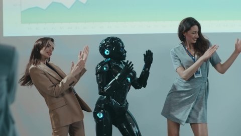 Beautiful happy women dancing with humanoid cyborg on robot presentation exhibition conference. Humans and robots. Funny trio. Celebration. Entertainment.