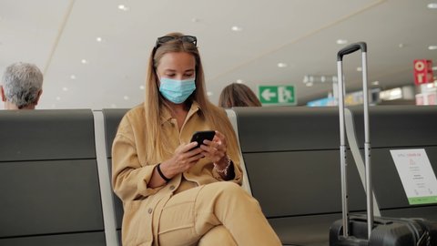 Woman tourist wearing medical protection mask using mobile phone in airport terminal, airline flight status and sit social distancing chair in airport during coronavirus post covid-19 virus outbreak