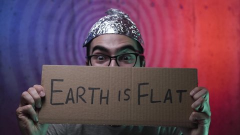 Strange young man wearing a tinfoil hat showing a cardboard sign with EARTH IS FLAT written on it. Weird men going insane about a conspiracy theory.