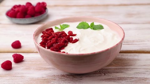 Male hand decorates yogurt with raspberry and goji in ceramic bowl on white wooden background. Side view, close up.