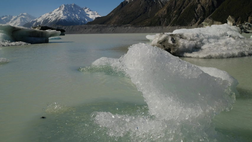 Icebergs floating on a beautiful lake and slowly melting in the sun with snow covered mountains and blue sky in the background. Located at Tasman Glacier river at Mt Cook in New Zealand.