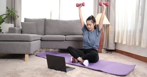 Sporty young asian woman doing exercising with dumbbells and watching fitness video tutorial online on laptop in the living room, Sport at home training online fitness class, Healthy lifestyle.
