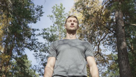 Good-looking muscular man in gray shirt standing still among trees with yellowing leaves looking thoughtfully at the distance. Low angle 4K 360 degree tracking arc shot.