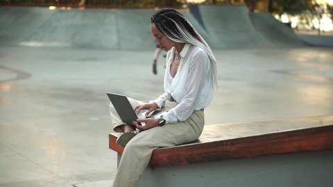 Side view of beautiful, elegant girl with black and white dreadlocks sitting on parapet in local skate park working on laptop, typing. Wearing nice white blouse and beige pants, Bike rider on Stock Video