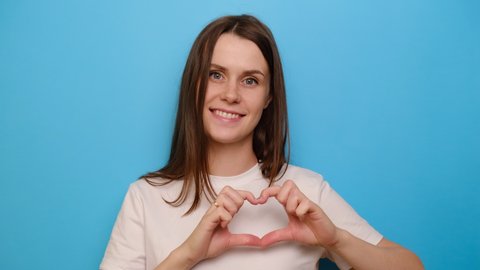 Young girl having wide charming smile put hands on chest make with fingers heart shape symbol of charity and human kindness, isolated on blue studio background. Generosity liberality sign concept