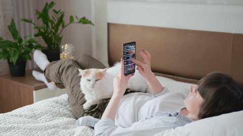 food delivery, young female shopper uses an app on smartphone and orders groceries from a store lying at home on bed with cat during isolation at lockdown due to pandemic and coronavirus, social