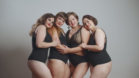 Group of happy oversize women in black bodysuits standing together over grey background. Four smiling plus size models posing at camera isolated. Body positive concept: stockvideo