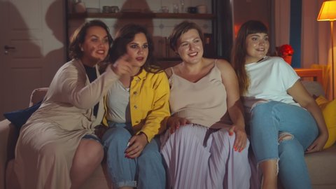 Smiling young oversize women relaxing and watching TV at home. Four fat girlfriends at slumber party watching movie in evening sitting on couch in living room. Friendship and body positive concept
